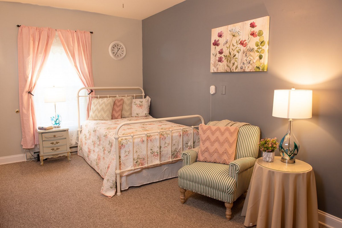 A cozy bedroom featuring a floral-themed decor with pastel colors, including a metal bed frame, patterned bedding, an armchair, and a soft lamp.