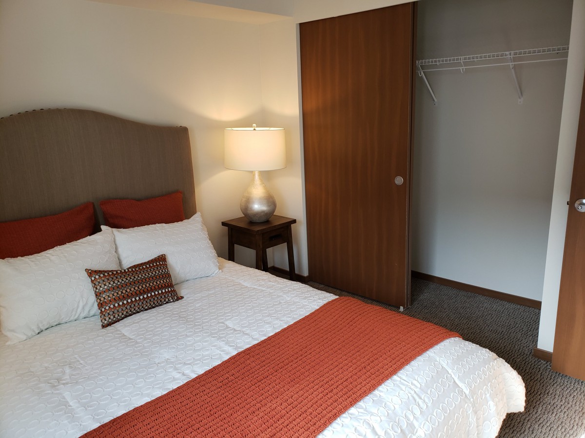 A cozy bedroom featuring a neatly made bed with orange accents, a bedside lamp, and an open closet.