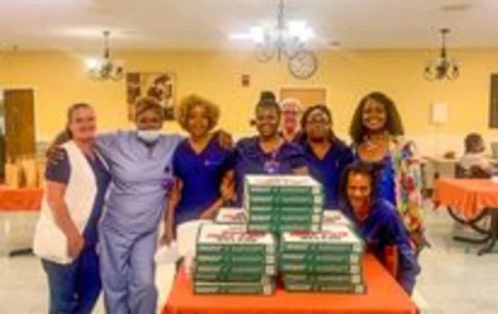 A group of healthcare professionals in uniform gathered around a table with stacks of pizza boxes.