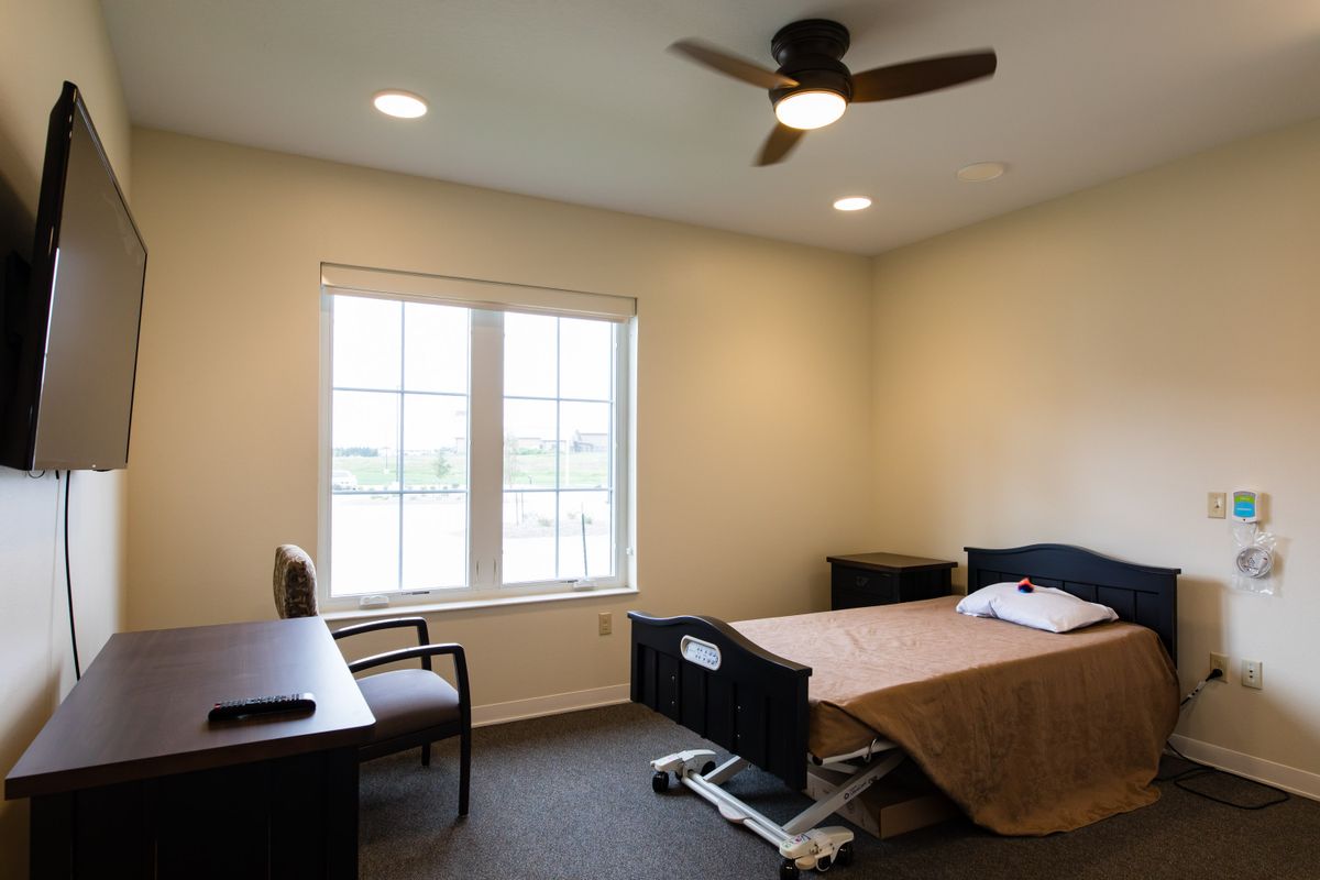 A simple, minimalist room with a hospital bed, desk, and chair under a ceiling fan, illuminated by natural light from a large window.