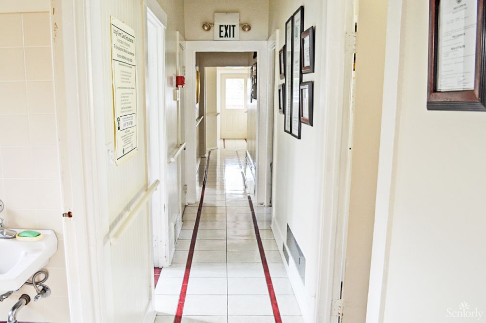 A bright hallway with tiled flooring, lined with doors and framed pictures, leading to an exit.