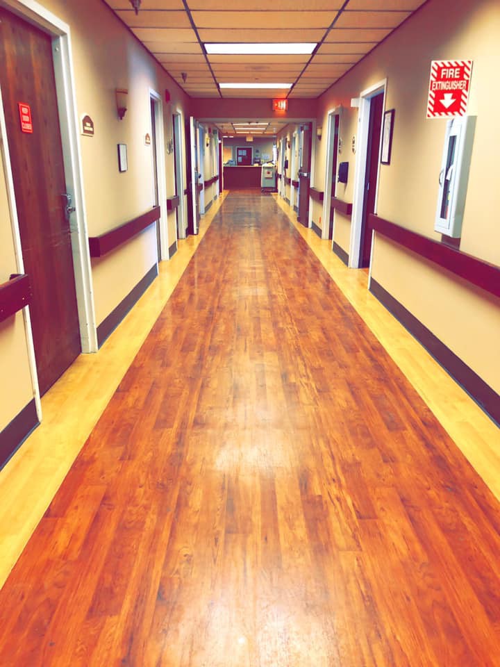 A long, well-lit hospital corridor with wooden floors and multiple patient rooms on either side.