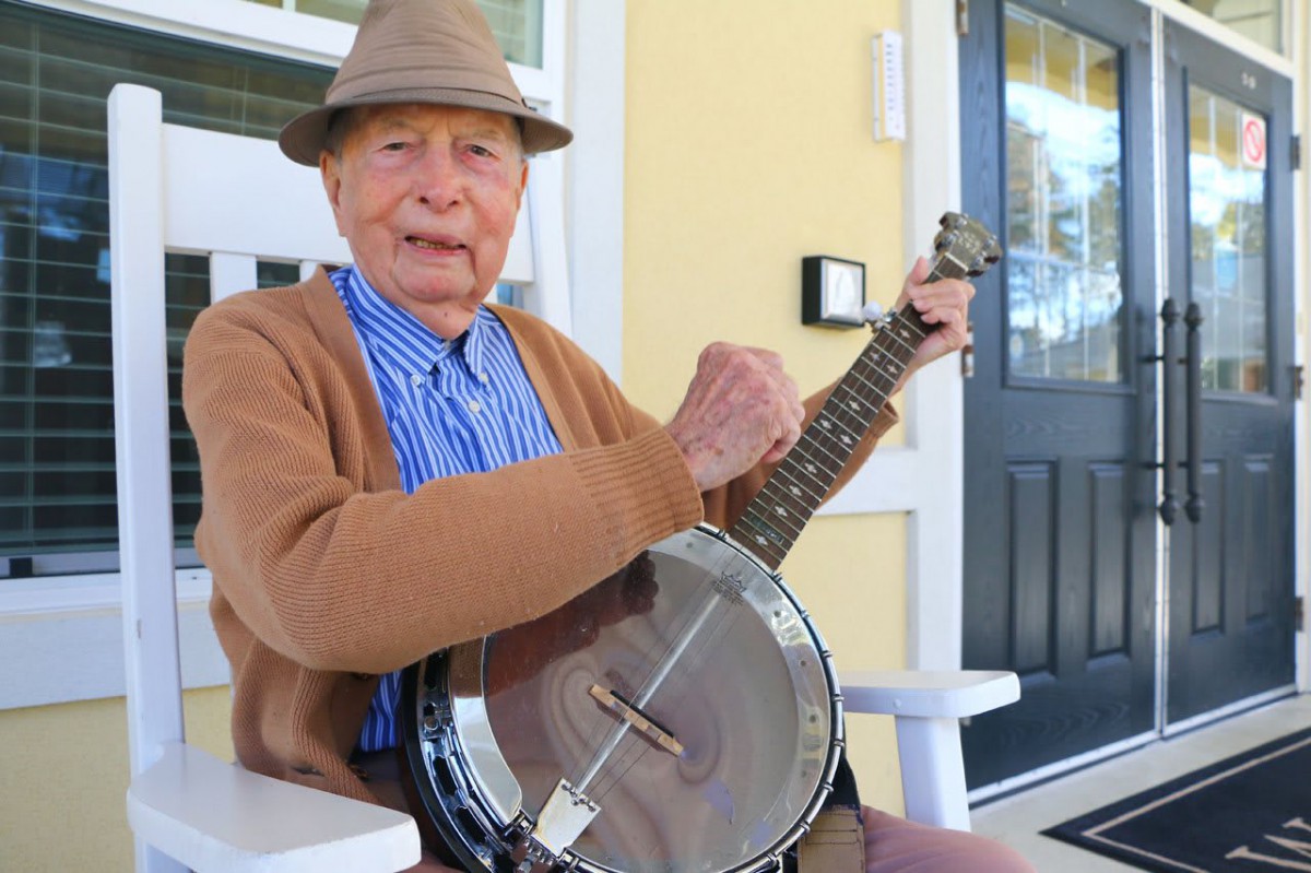 An elderly man playing a banjo while sitting on a porch.