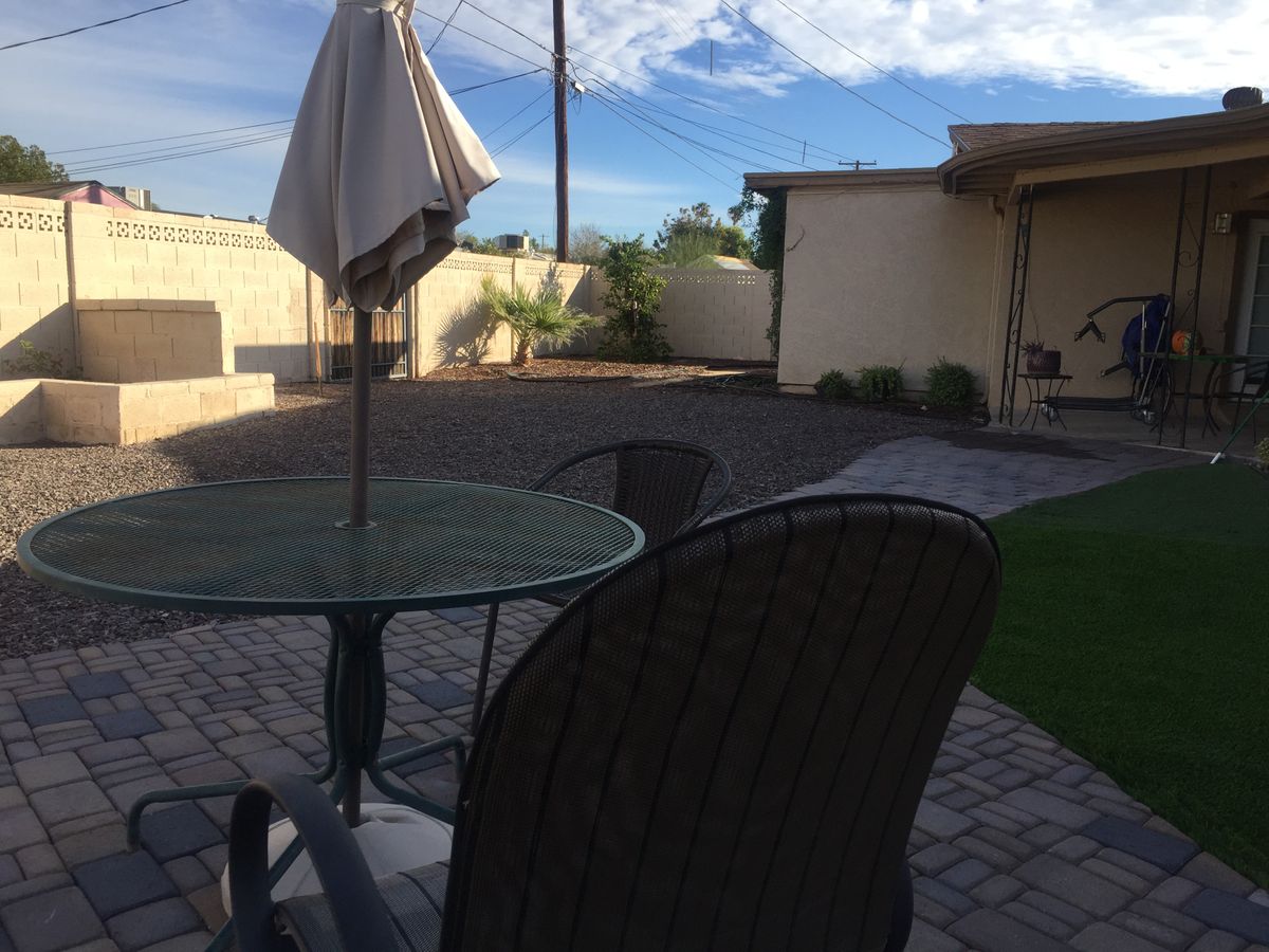 A backyard patio with a glass table, chairs, and an umbrella, set on a paved area under a sunny sky.