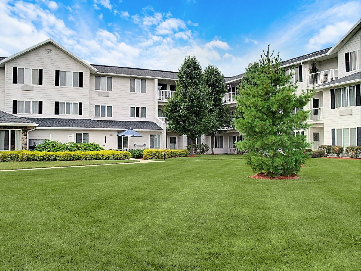 A residential apartment building with a well-maintained lawn and surrounding trees under a blue sky.