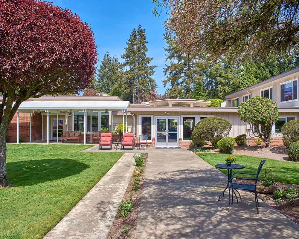 A serene backyard view of a residential home with a patio, lawn, and outdoor seating surrounded by trees.