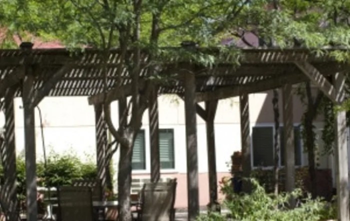 A peaceful garden setting featuring a pergola, trees, and outdoor furniture under dappled sunlight.