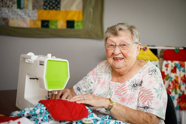 An elderly woman is happily engaged in sewing, working on a colorful quilt with a sewing machine.