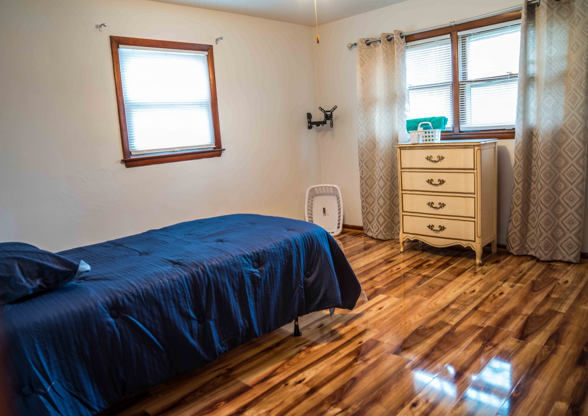 A cozy bedroom with wooden flooring, featuring a bed with a blue bedspread and a dresser by the window.