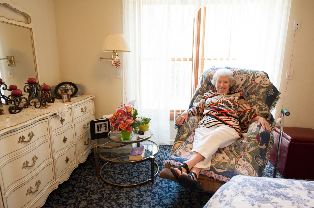An elderly woman relaxing in a cozy living room with floral decor and bright natural light.