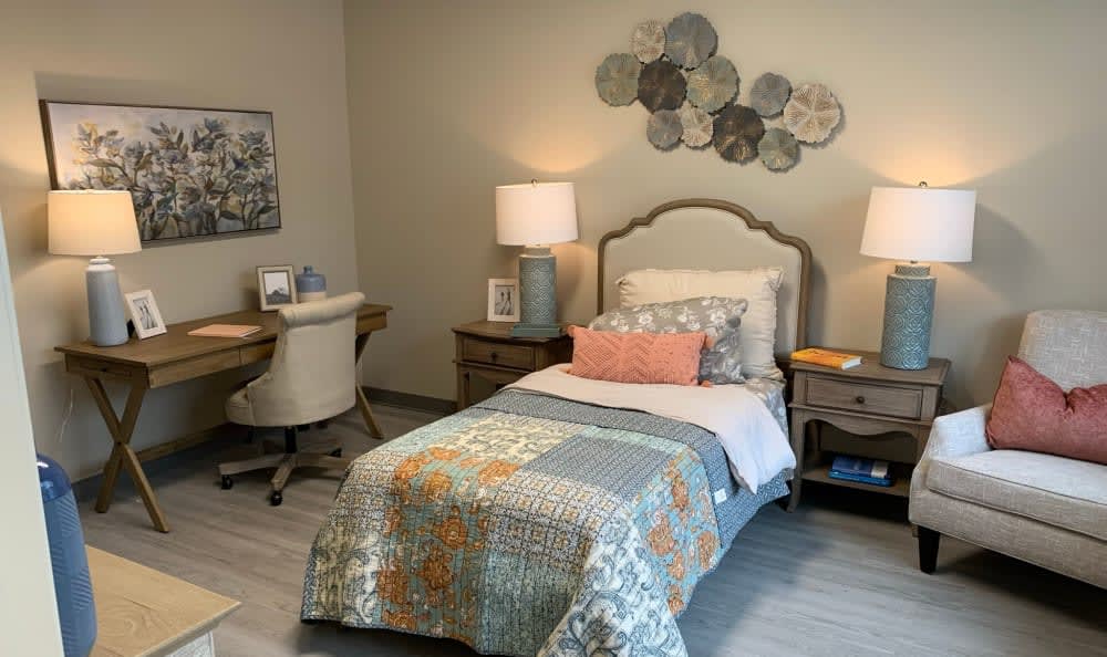 A cozy bedroom featuring a single bed with decorative pillows, accompanied by a desk and chair, illuminated by table lamps, and adorned with wall decor.