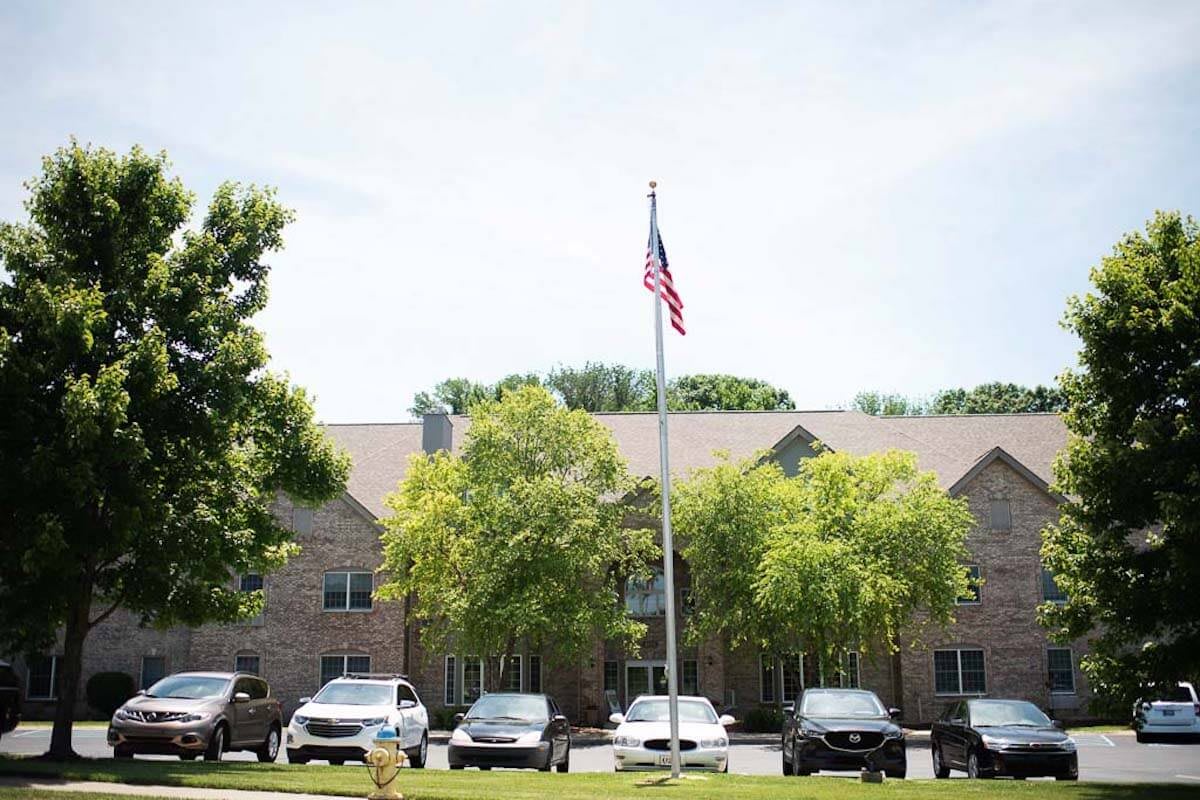 A building with a US flag in front, surrounded by parked cars and trees.