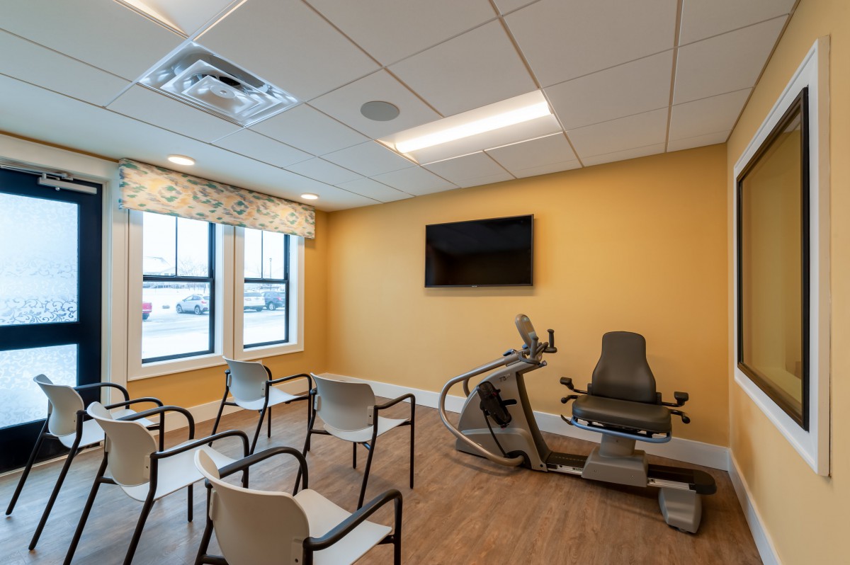 A well-lit fitness room with chairs arranged facing a TV and an exercise machine.