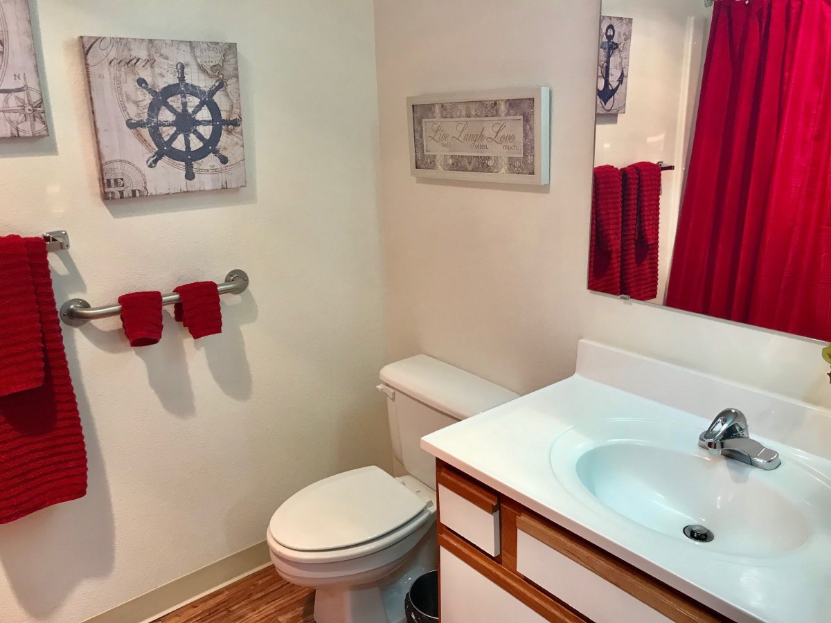 A small bathroom with nautical-themed decor, featuring a sink, toilet, mirror, and red towels.