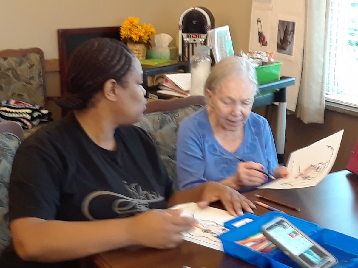 An elderly woman is engaged in a drawing activity with the assistance of a caregiver.