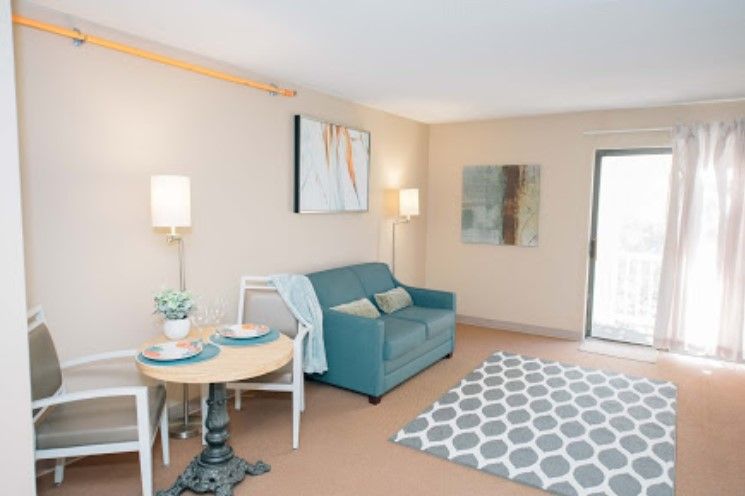 A cozy living room with a teal sofa, artwork on the walls, a small dining table, and a patterned rug.