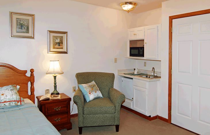 A cozy living space with a chair, lamp, and small kitchen area.