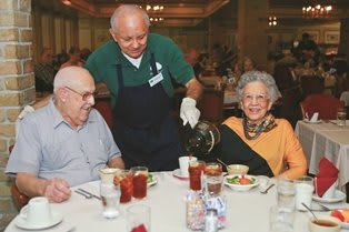An elderly couple enjoys a meal at a restaurant while being served by a waiter.
