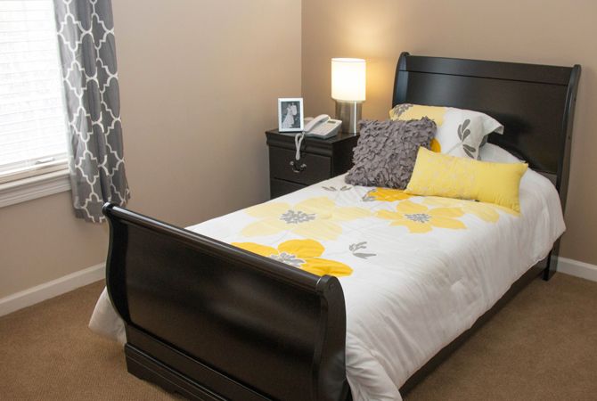A cozy bedroom with a single bed, adorned with decorative pillows and a floral blanket. A lamp and photo frame sit on the nightstand.