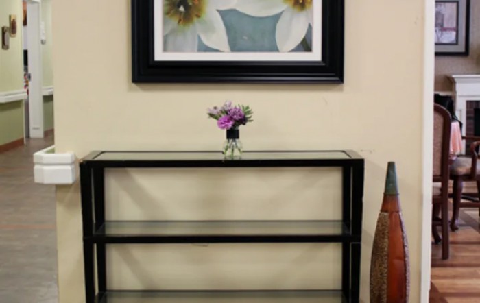 A minimalist glass shelf with a small vase of flowers, situated beneath a framed floral artwork.
