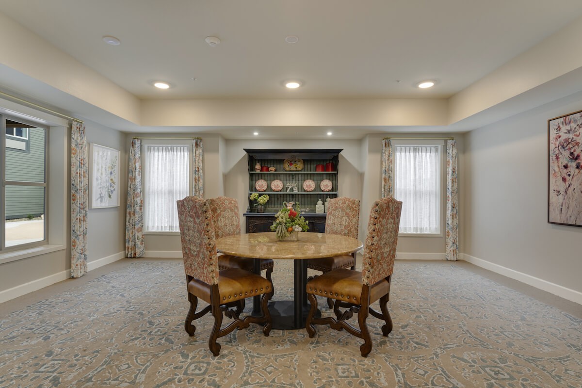 A cozy dining room features a round table surrounded by four upholstered chairs, with a shelving unit in the background.