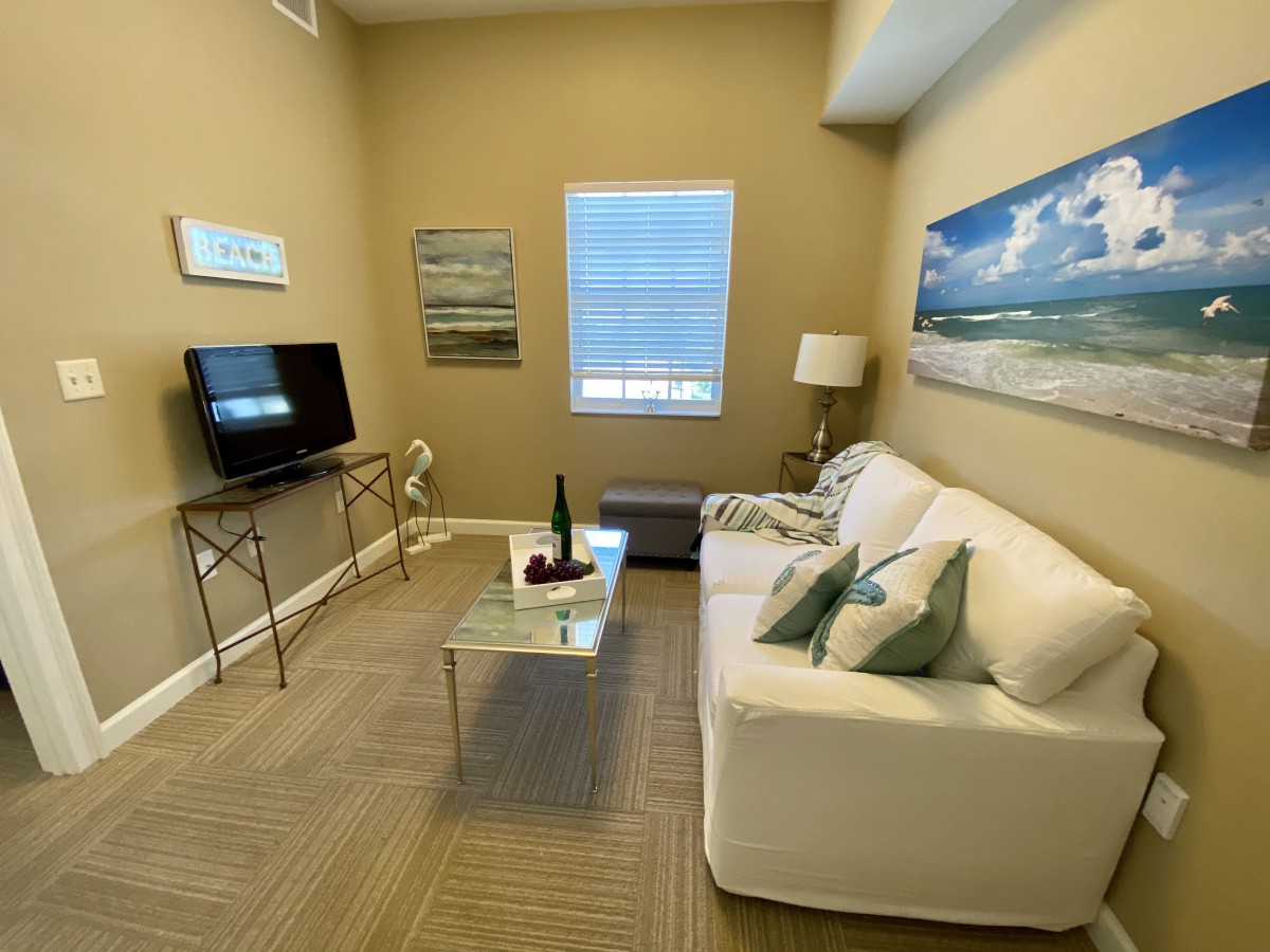 A cozy living room with beige walls, featuring a white sofa, a glass coffee table, and a television on a small console.
