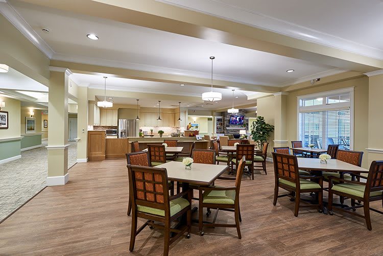 A well-lit dining area featuring multiple tables and chairs, with a view of the kitchen in the background.