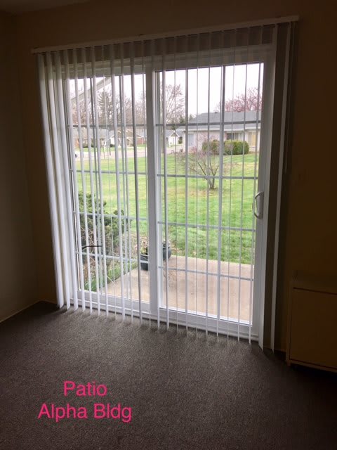 A room with a sliding glass door and vertical blinds, overlooking a patio and grassy yard.