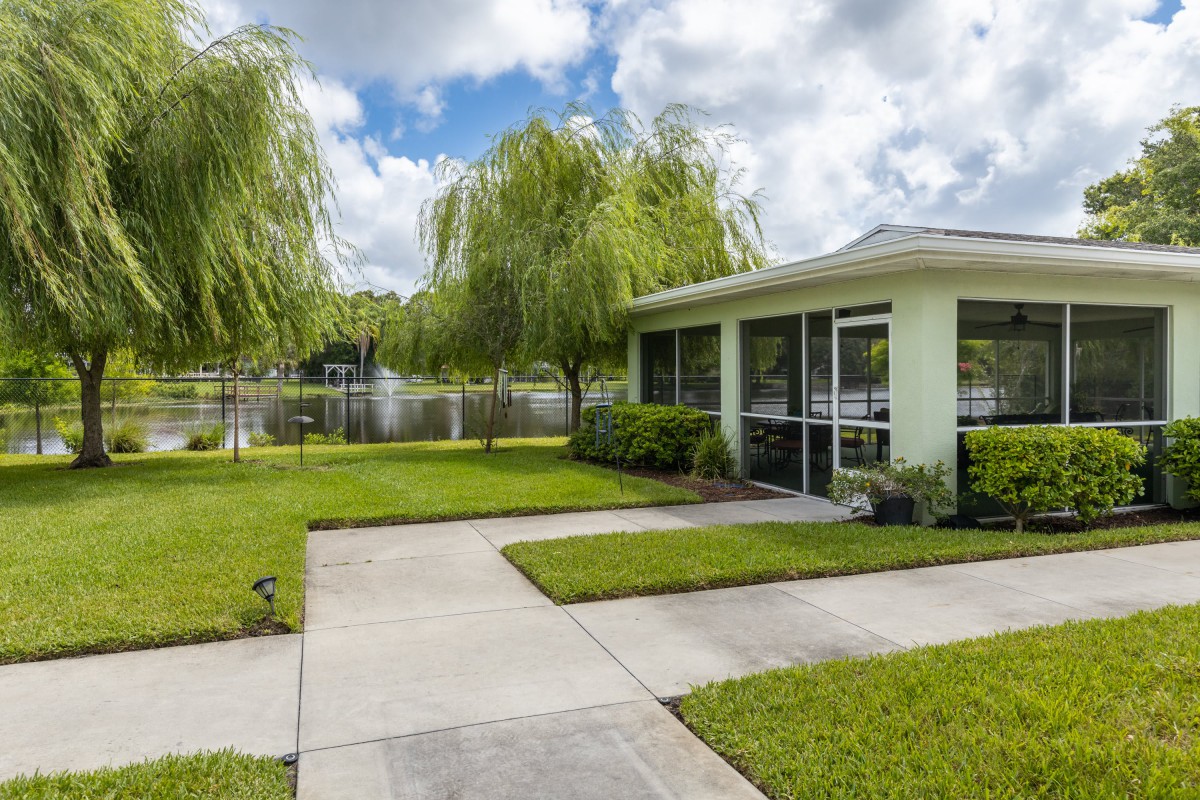 A modern house with a green lawn and driveway, situated next to a lake under a cloudy sky.