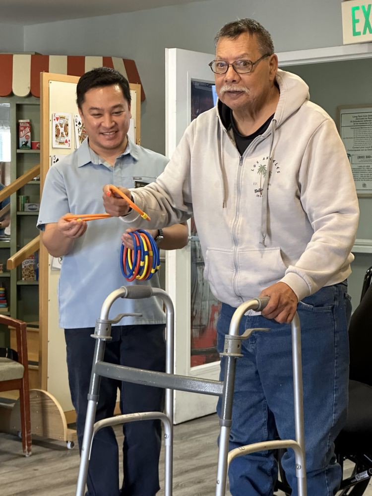 An elderly man using a walker for support during a physical therapy session indoors, assisted by a caregiver.