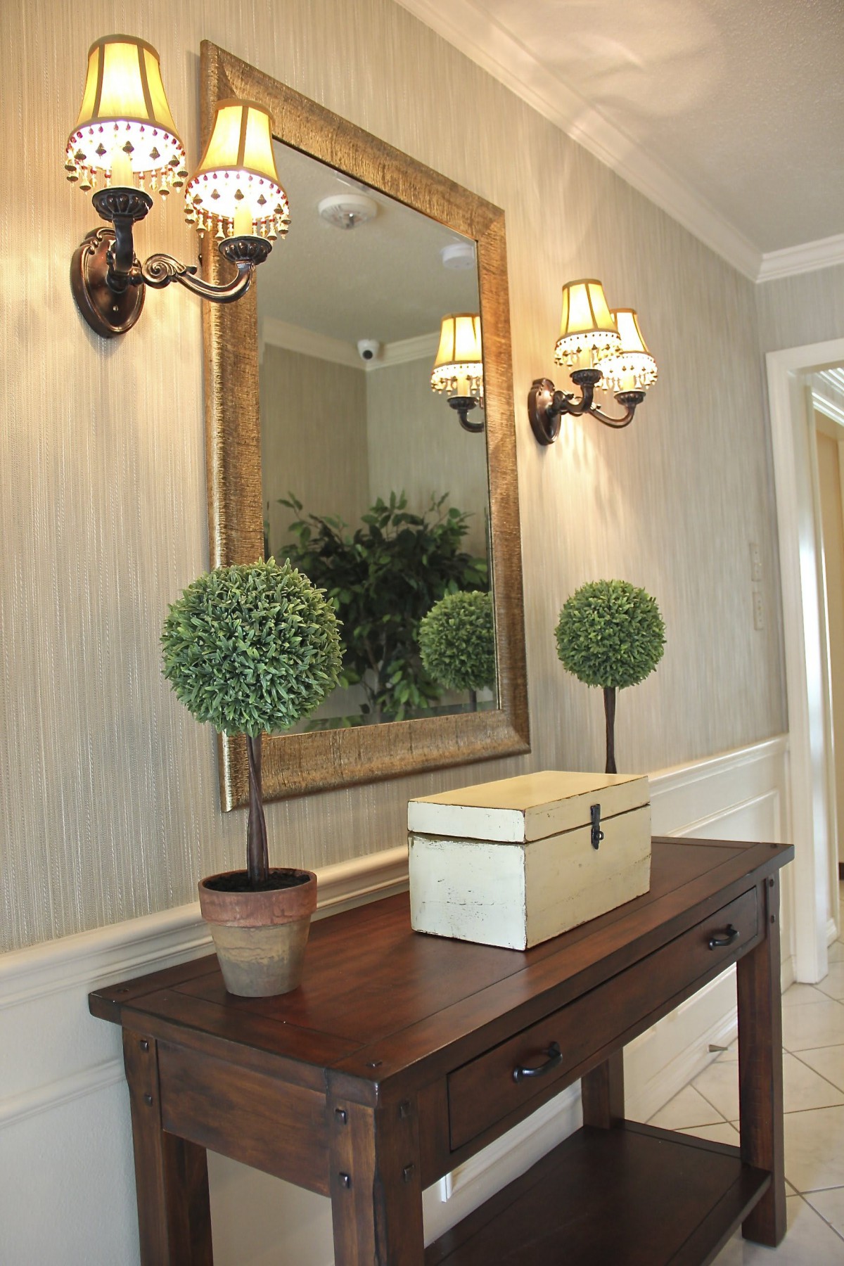 A stylish hallway features a wooden console table, decorative potted plants, and an ornate mirror with elegant wall sconces.