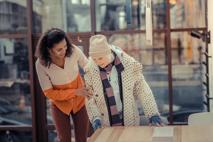 A young woman assists an elderly lady indoors, providing care and support.