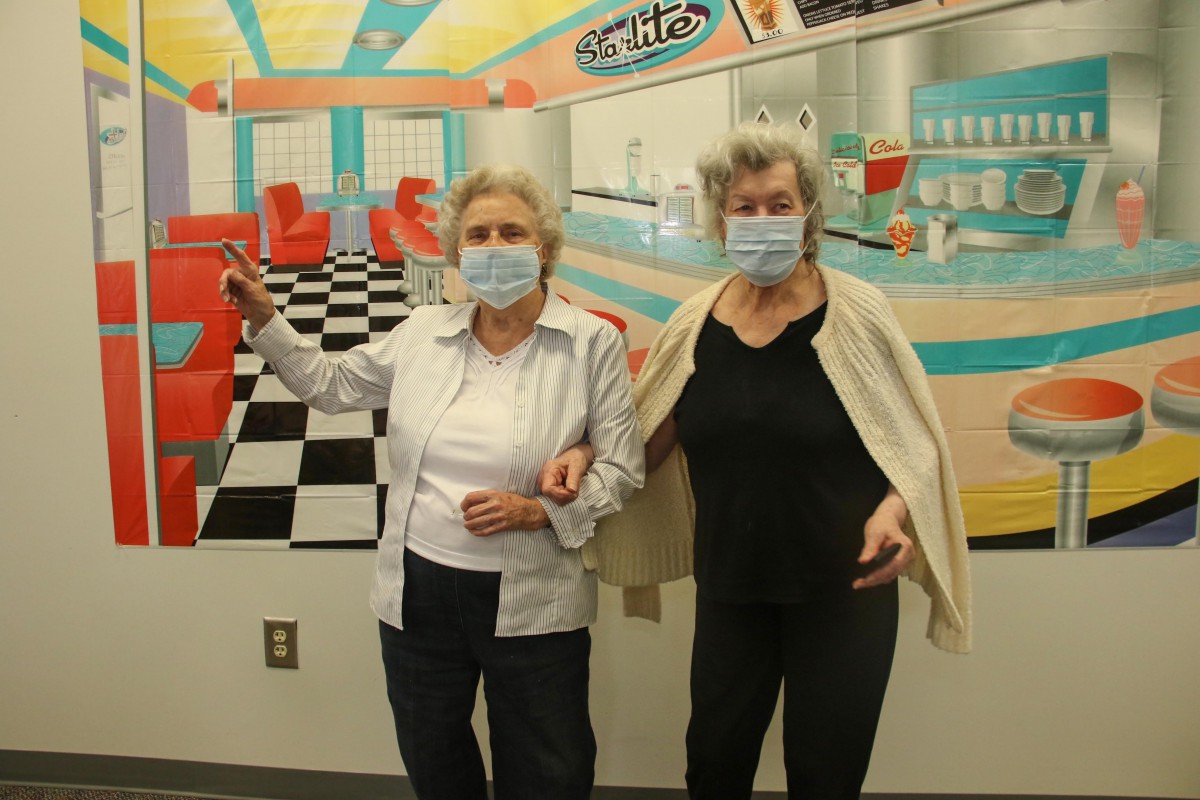 Two people wearing masks stand in front of a retro diner-themed backdrop.