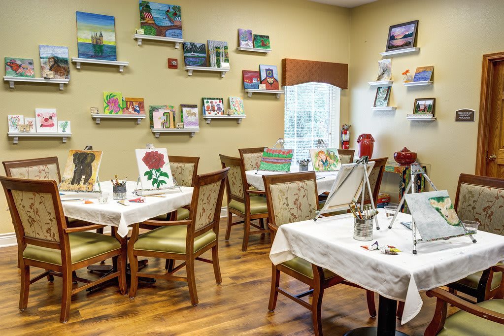 An art studio with tables set up for painting, featuring canvases on easels and completed artwork displayed on the walls.