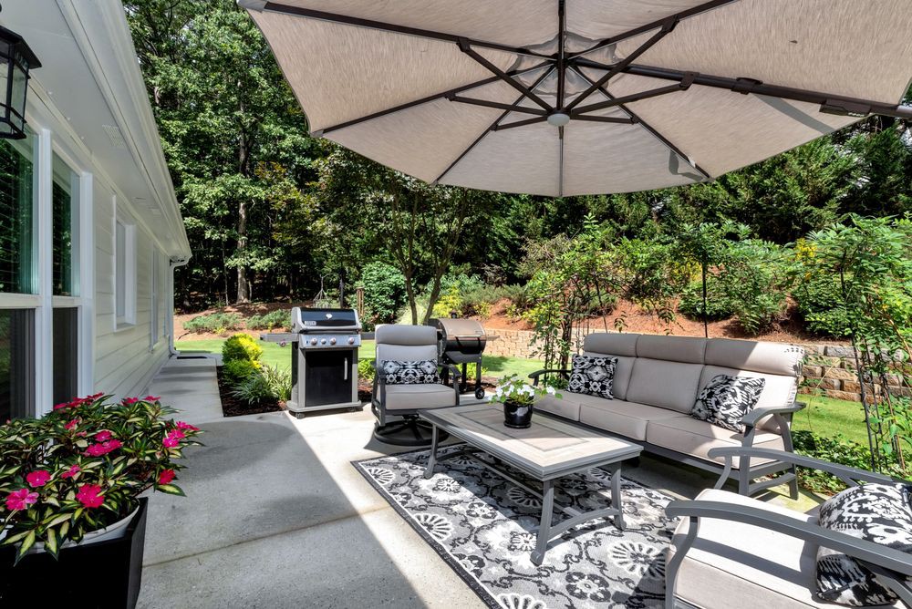 A cozy outdoor patio with comfortable seating, a large umbrella, and a grill, surrounded by lush greenery.