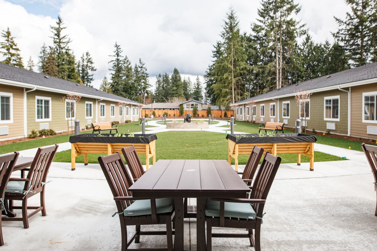 A scenic outdoor patio area with a wooden table and chairs, surrounded by benches, buildings, and evergreen trees under a cloudy sky.