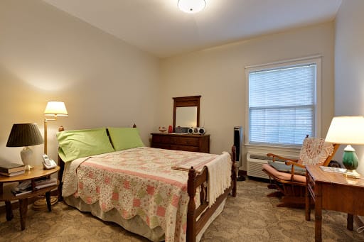 A cozy bedroom featuring a bed with a quilt, a dresser with a mirror, and a comfortable chair next to a window.