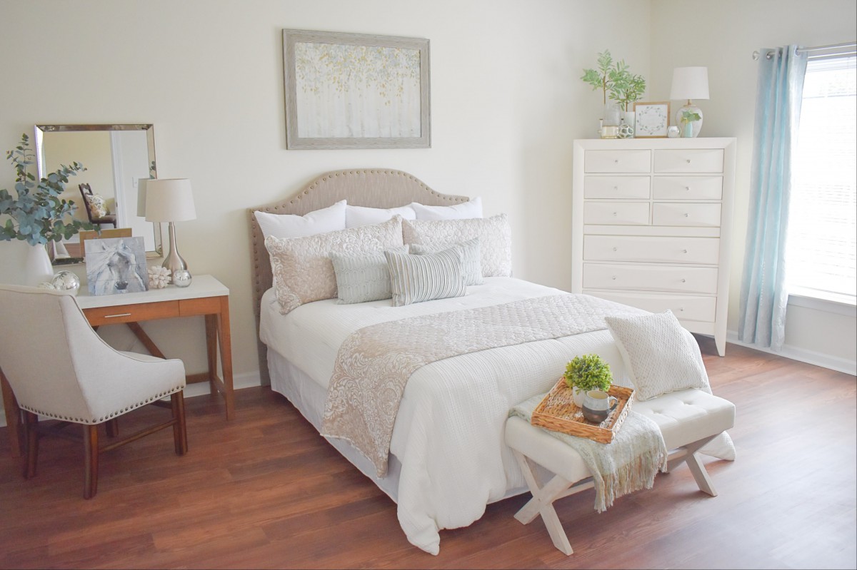A cozy, minimalist bedroom featuring a neatly made bed with light-colored bedding and decorative pillows, a dresser, and soft lighting.