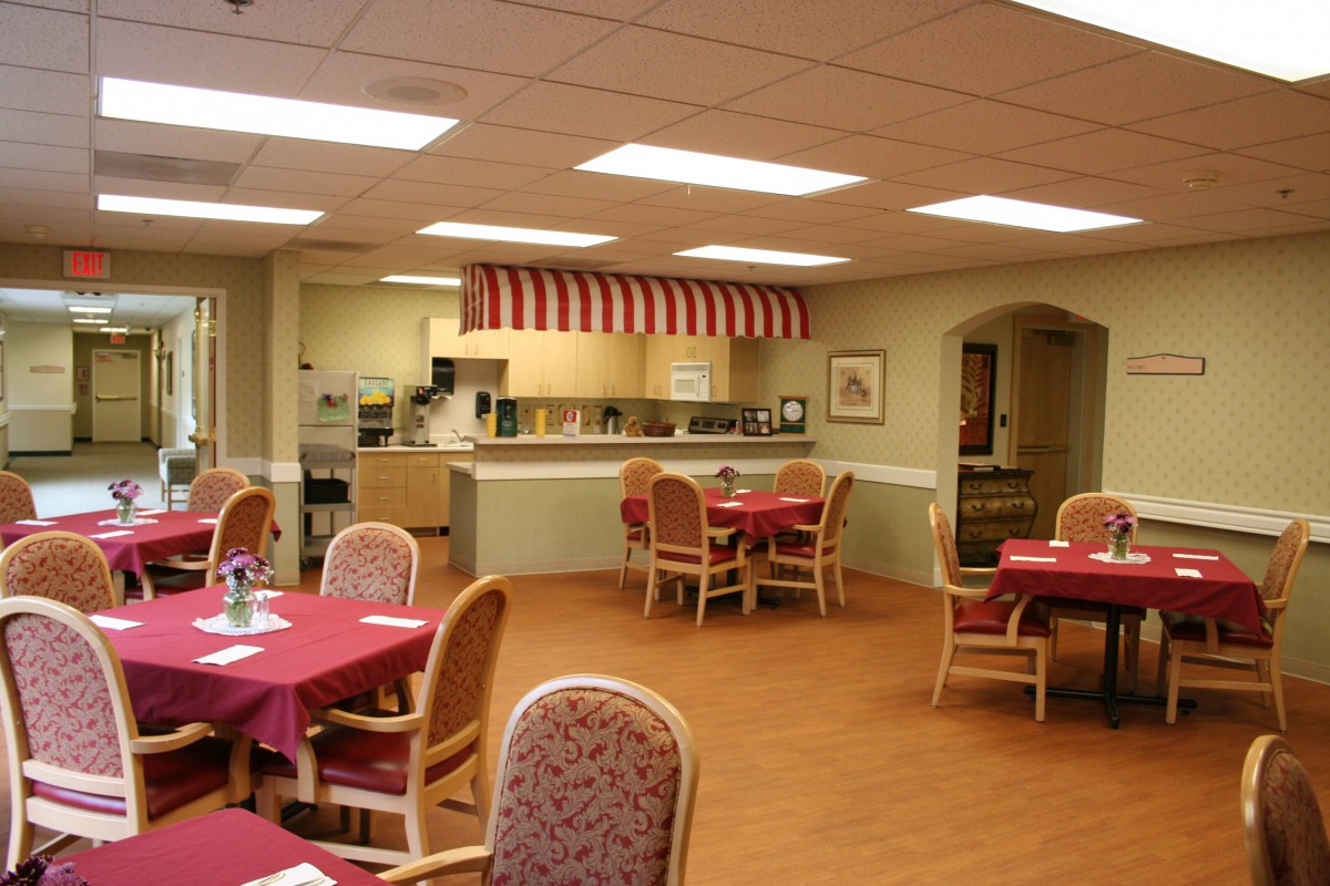A cozy dining area featuring neatly arranged tables with red tablecloths and a small open kitchen in the background.