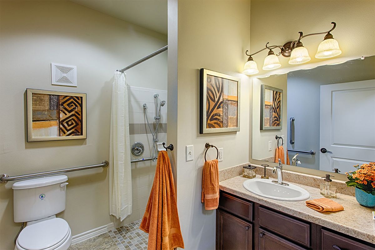 A neatly decorated bathroom with a shower, sink, and mirror, adorned with orange towels and artwork.