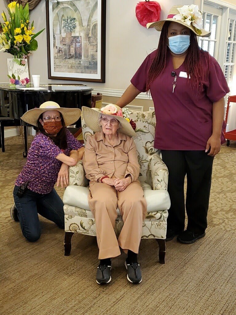 An elderly person seated in a chair with two caregivers, all wearing hats, inside a care facility.