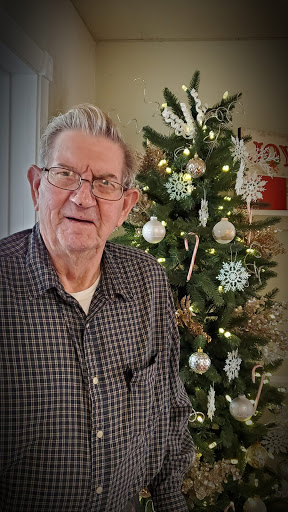 An elderly man poses cheerfully in front of a beautifully decorated Christmas tree.