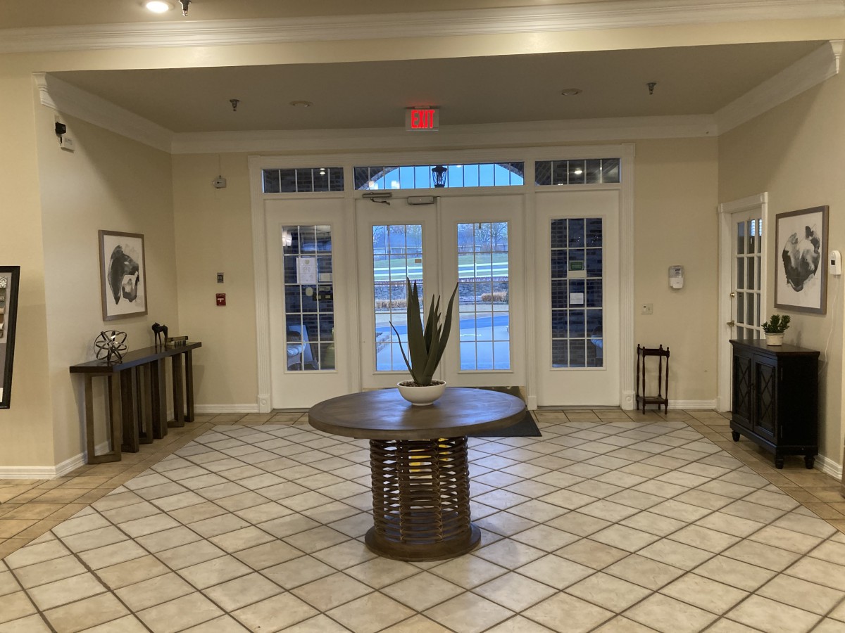 A spacious lobby area with tiled flooring, featuring a central table adorned with a plant and large windows providing natural light.