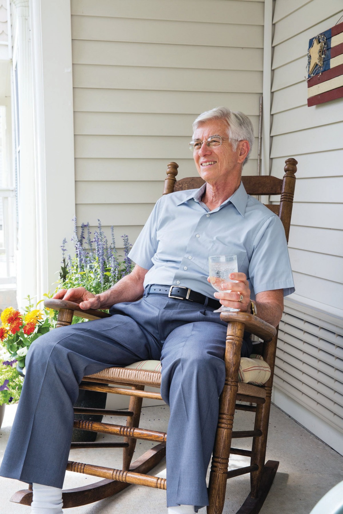An elderly man relaxing in a rocking chair on a porch, holding a glass and surrounded by flowers.