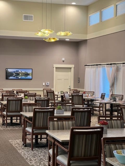 An empty dining room with neatly arranged tables and chairs, featuring a wall-mounted television and elegant lighting.