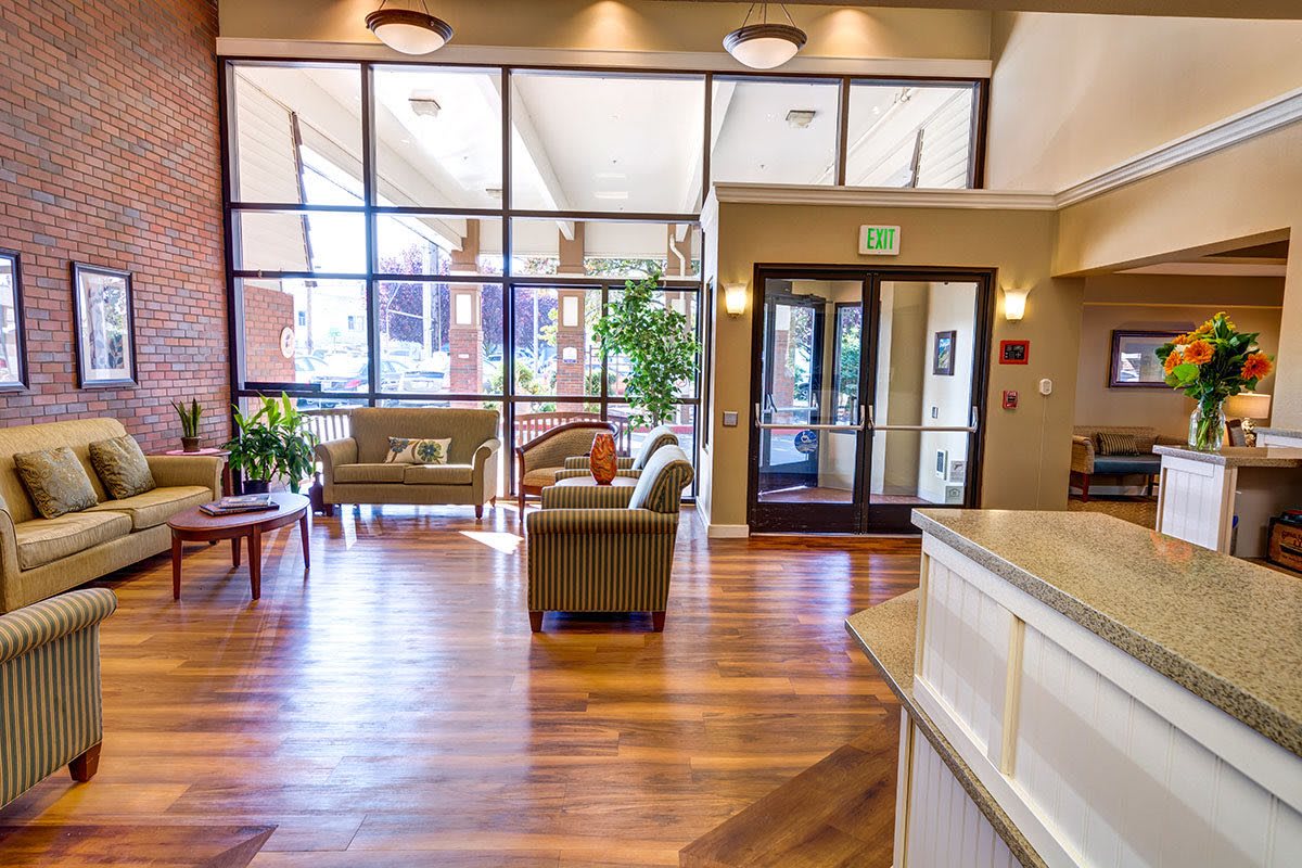 A bright and inviting lobby with a spacious waiting area, featuring comfortable seating, wooden flooring, large windows, and decorative plants.