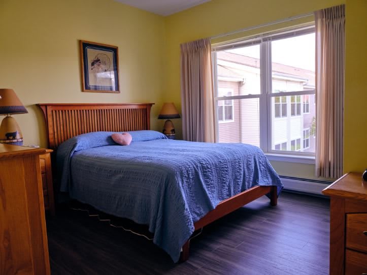 A cozy bedroom with a wooden bed frame, blue bedding, and large windows letting in natural light.