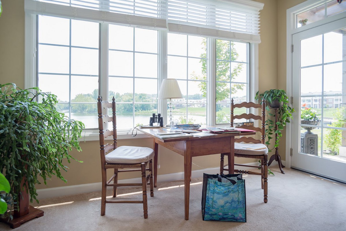 A cozy sunroom with a wooden table and chairs, filled with natural light from large windows and decorated with plants.