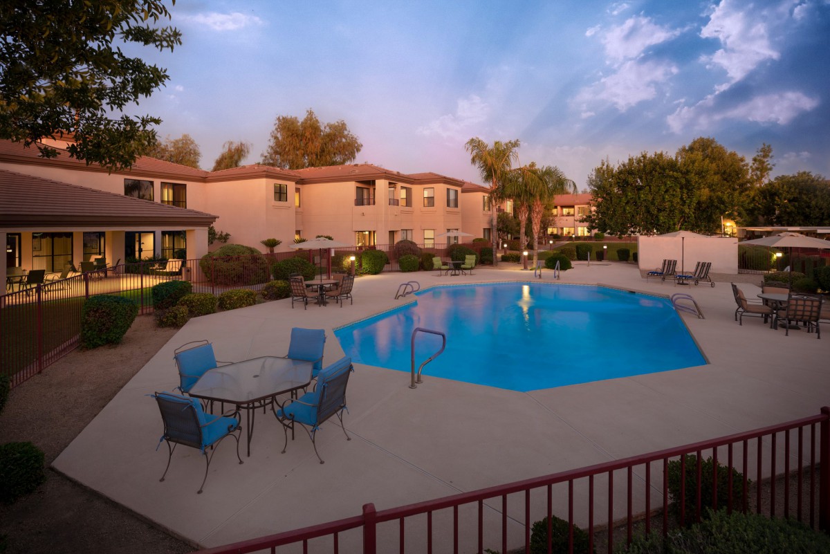A serene outdoor view of a residential swimming pool surrounded by trees and patio furniture, located in an apartment complex.
