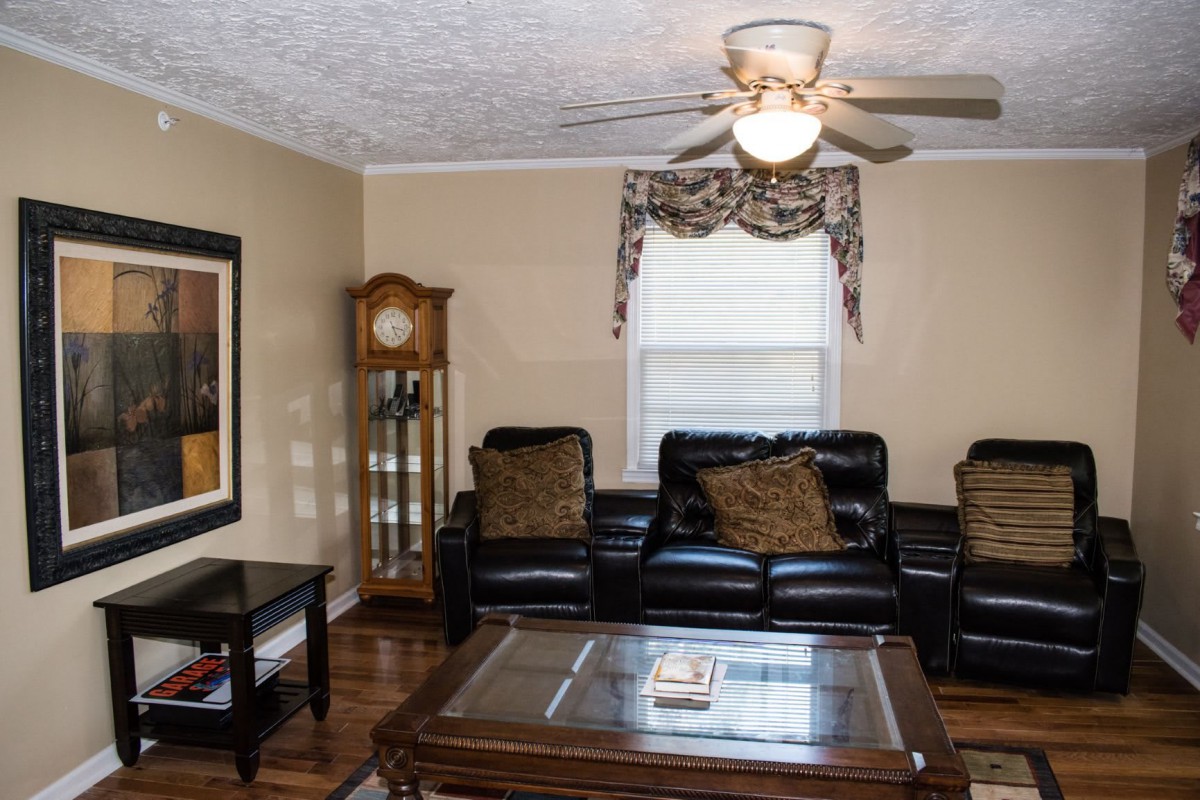 A cozy living room with a leather sofa, glass-top coffee table, grandfather clock, and ceiling fan.